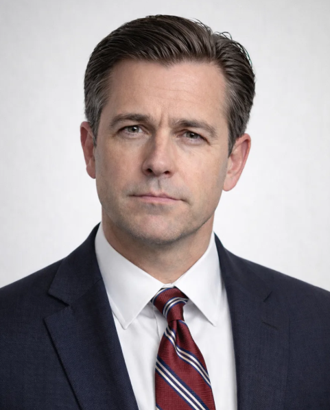 Professional headshot of a man in a dark suit, white shirt, and striped tie against a neutral gray background.